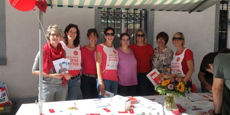 8 Frauen stehen fröhlich draußen an einem Infostand für den Blutspendedienst