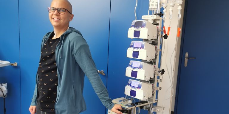 Myriam steht im Spital, angeschlossen an einen Infusionsständer. Die Haare sind ihr von der Chemotherapie bereits ausgefallen.