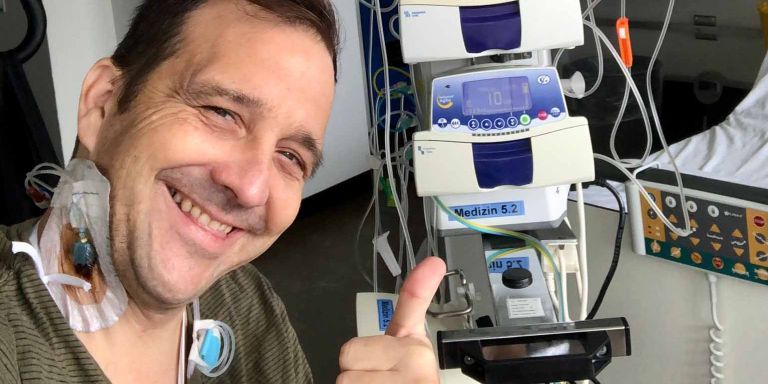 Christian Bregy blickt lächelnd in die Kamera. Er hat einen Halsvenenkatheter und im Hintergrund sind die Geräte zu sehen, die für seine Chemotherapie benötigt werden.