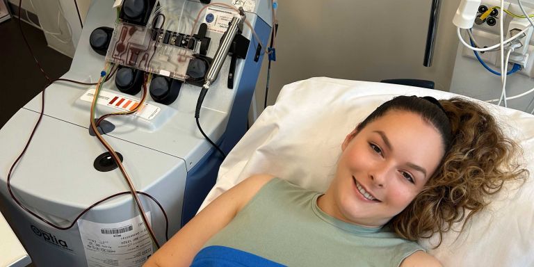 Christine, Blutstammzellspenderin, liegt im Spitalbett und lächelt in die Kamera. Neben ihr ist das Apharesegerät zu sehen.