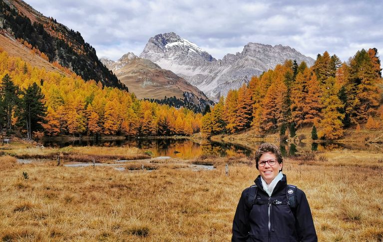 Myriam steht vor einer Engadiner Herbstlandschaft. Im Hintergrund sieht man Tannen und Berge. 