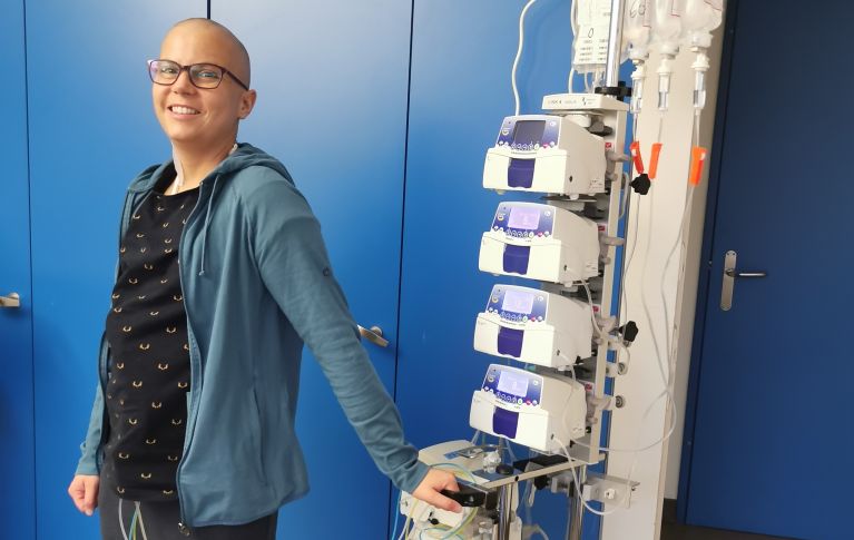 Myriam steht im Spital, angeschlossen an einen Infusionsständer. Die Haare sind ihr von der Chemotherapie bereits ausgefallen.