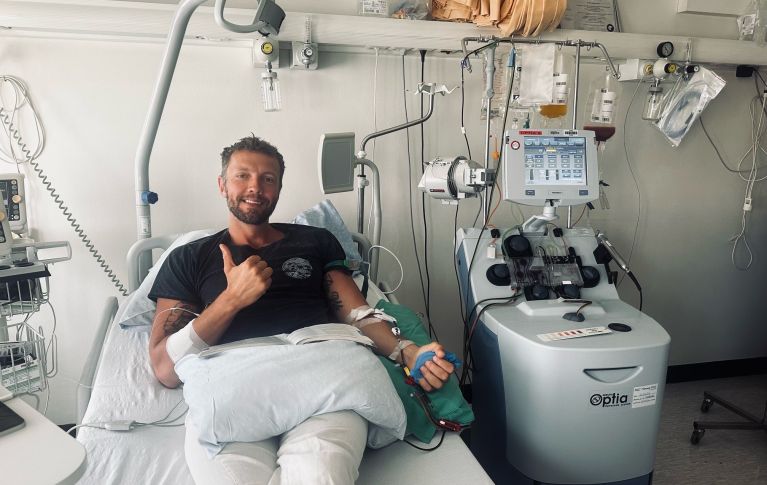 Il donatore di cellule staminali del sangue Damien G. è sdraiato su un letto d'ospedale. Ad entrambe le braccia sono collegati dei cateteri venosi che convogliano il sangue verso il cosiddetto dispositivo di afresi e di nuovo nel corpo. Damien fa il segno del pollice in su