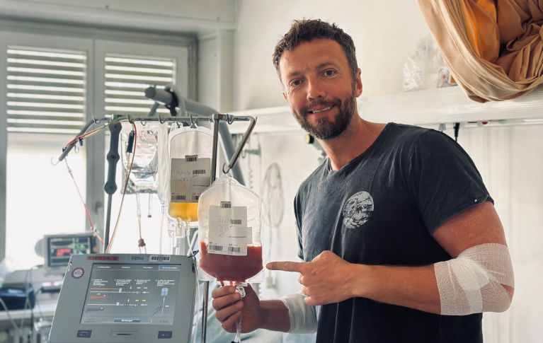 Il donatore di cellule staminali del sangue Damien G. sorride alla telecamera e indica la sacca contenente le sue cellule staminali del sangue donate.