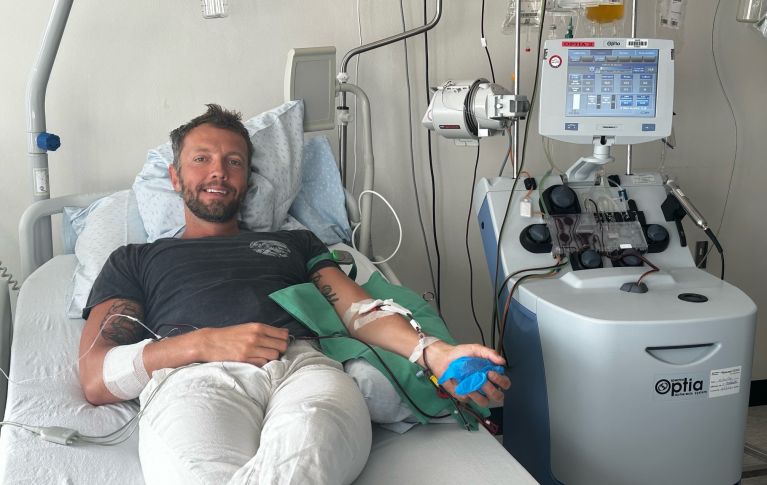 Il donatore di cellule staminali del sangue Damien G. è sdraiato su un letto d'ospedale. Ad entrambe le braccia sono collegati dei cateteri venosi che convogliano il sangue verso il cosiddetto dispositivo di afresi e di nuovo nel corpo. 