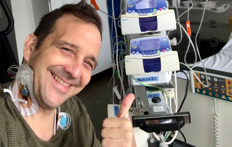 Christian Bregy guarda la telecamera con un sorriso. Ha un catetere nella vena giugulare e sullo sfondo si vedono le apparecchiature necessarie per la chemioterapia.