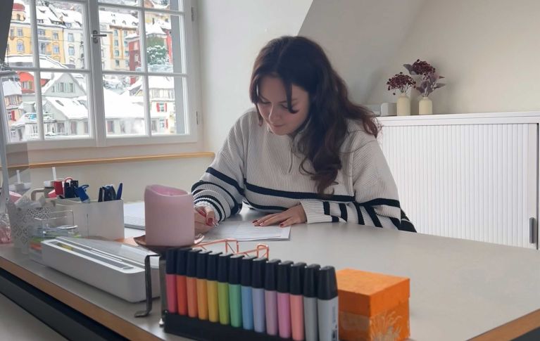 Christine, eine junge Frau mit lockigem, braunen Haar sitzt an einem Schreibtisch und korrigiert Schularbeiten.