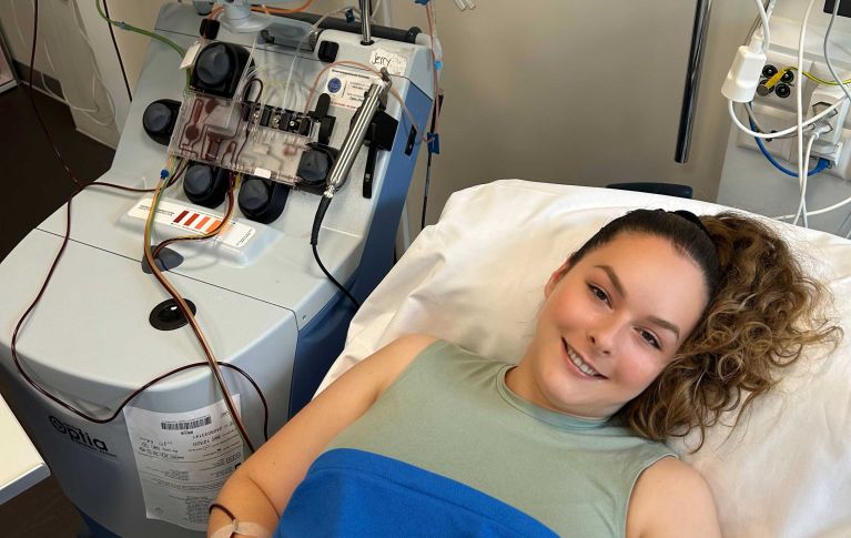 Christine, donatrice di cellule staminali del sangue, giace nel suo letto d'ospedale e sorride alla telecamera. Accanto a lei si vede la macchina per l'aparesi.