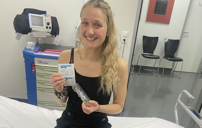 Marion, eine junge Blutstammzellspenderin, sitzt auf einer Spitalliege und hält die Spritze mit den Wachstumsfaktoren in die Kamera.