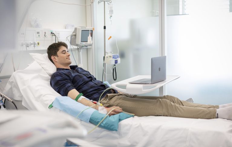 Ein junger Mann bei der peripheren Blutstammzellspende. Er in einem Spitalbett, an seinen Armen sind die venösen Zugänge sichtbar.