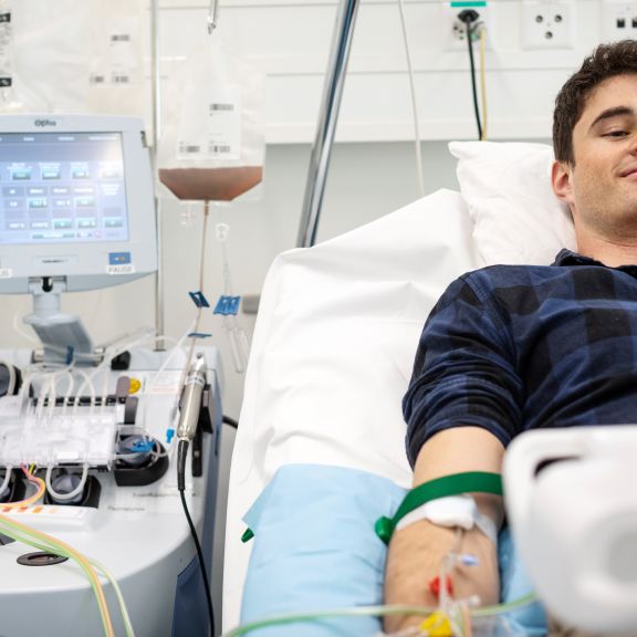 Ein junger Mann bei der peripheren Blutstammzellspende. Er liegt in einem Spitalbett und blickt auf einen Laptop.