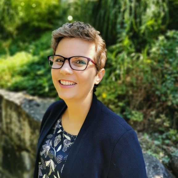 Porträtfoto von Myriam, sie lächelt in die Kamera und befindet sich draussen im Grünen