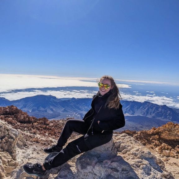 Ladina Gall en randonnée, assise sur un rocher, derrière elle une vue dégagée sur la vallée, ciel bleu.